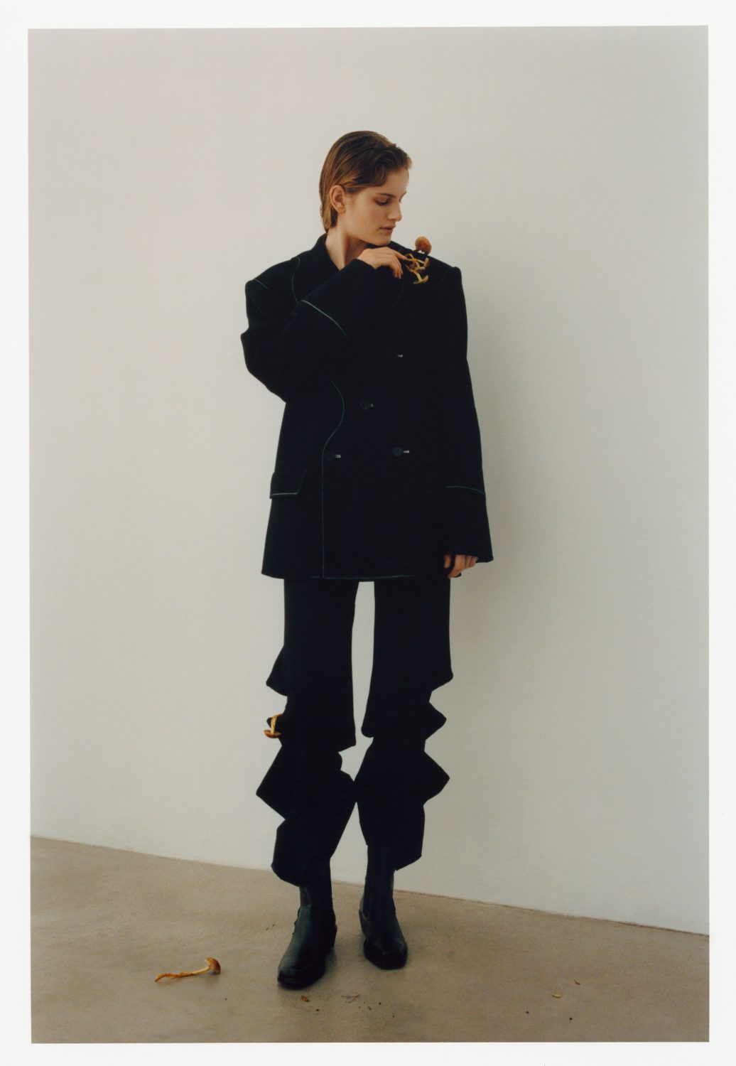 Fashionable young woman in oversized black suit with ruffles, standing indoors in front of a neutral wall and looking at jewelry.