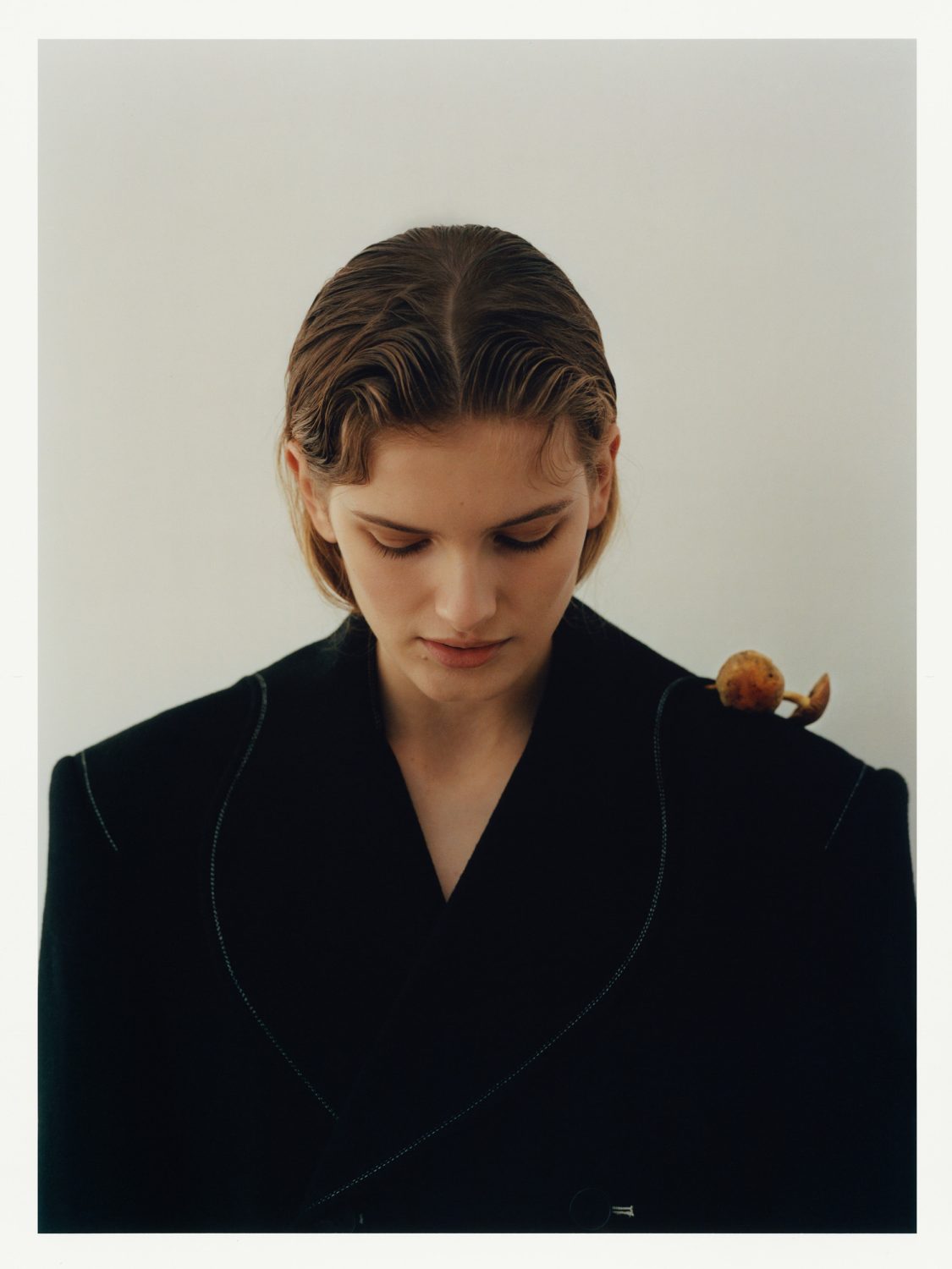 Close-up studio shot of a young fair-skinned woman with wet-look wavy hair wearing a dark business jacket with an orange fruit on her shoulder.