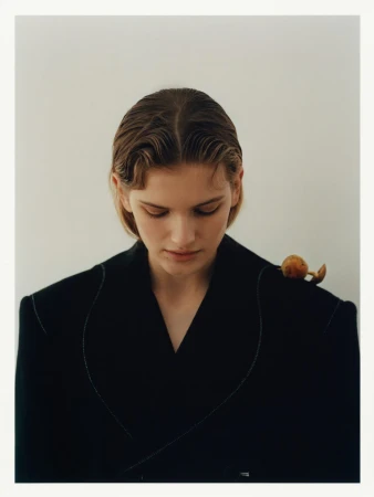 Close-up studio shot of a young fair-skinned woman with wet-look wavy hair wearing a dark business jacket with an orange fruit on her shoulder.