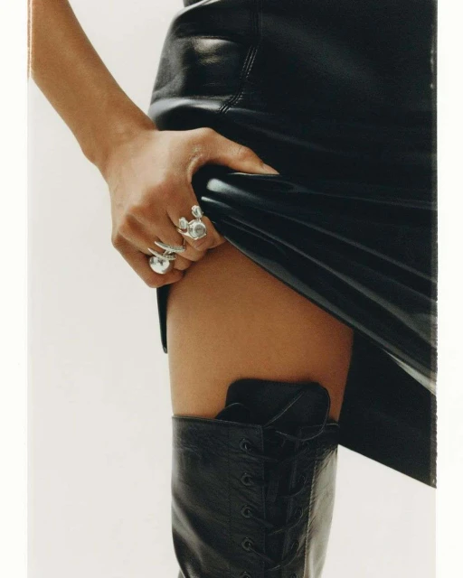 Close-up of a Black leather mini skirt and lace-up boots worn by an adult female, accessorized with statement rings on her left hand.