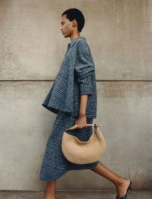 An adult woman with short dark hair walking in a grey textured matching set and carrying a crescent-shaped handbag outdoors.