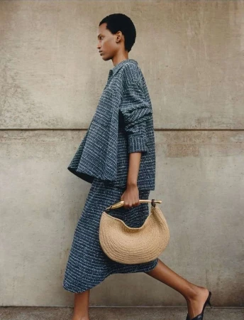 An adult woman with short dark hair walking in a grey textured matching set and carrying a crescent-shaped handbag outdoors.