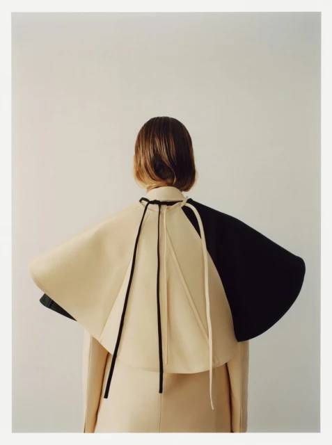 Back view of a female in a beige outfit with a two-tone black and beige cape. Neutral studio background. Fashion editorial shot.