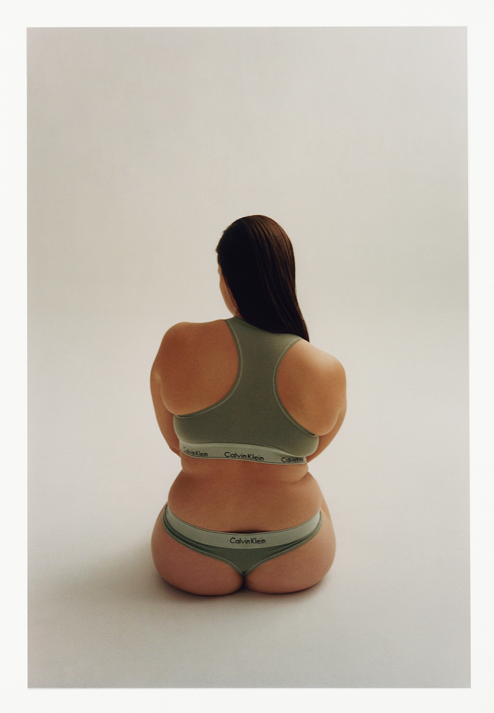 Back view of an adult female sitting in a photo studio, wearing matching green Calvin Klein underwear and sports bra against a cream background.