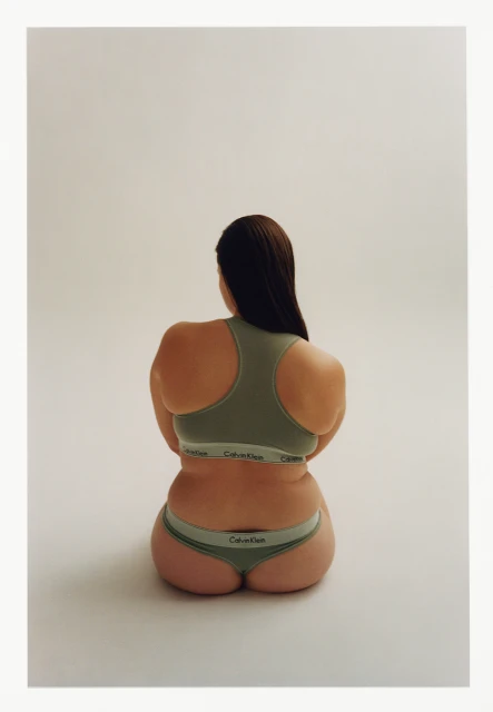 Back view of an adult female sitting in a photo studio, wearing matching green Calvin Klein underwear and sports bra against a cream background.