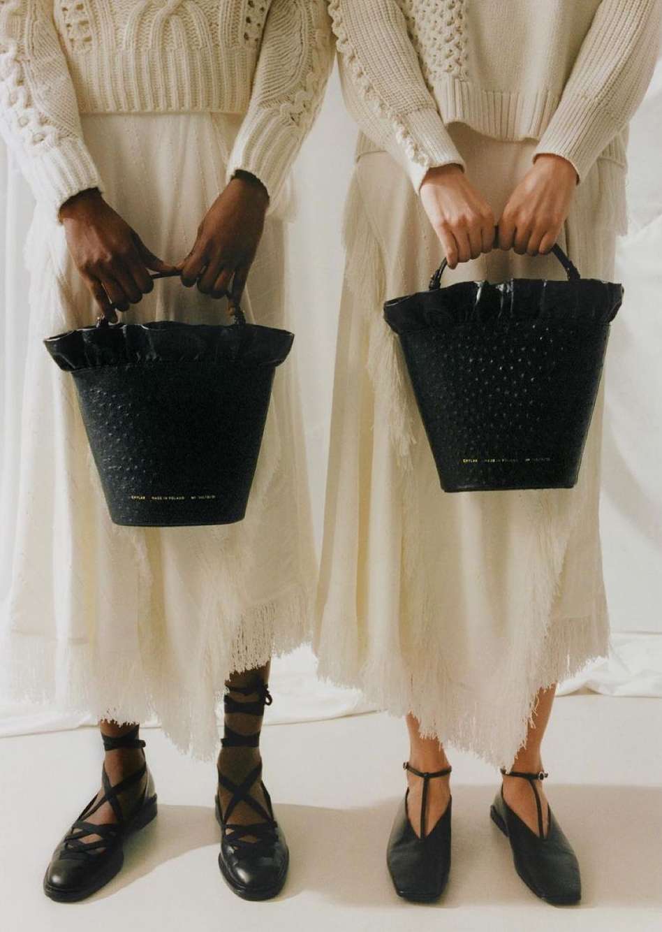 Two women in white dresses and black flats hold black woven bags against a white backdrop in a fashion or editorial image.