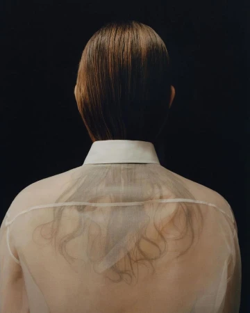 Rear view of a person wearing a sheer white button-down shirt with straight brown hair falling down the back against a dark background