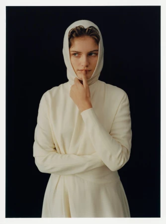 Model wearing a white hooded top with finger on lips against a black background