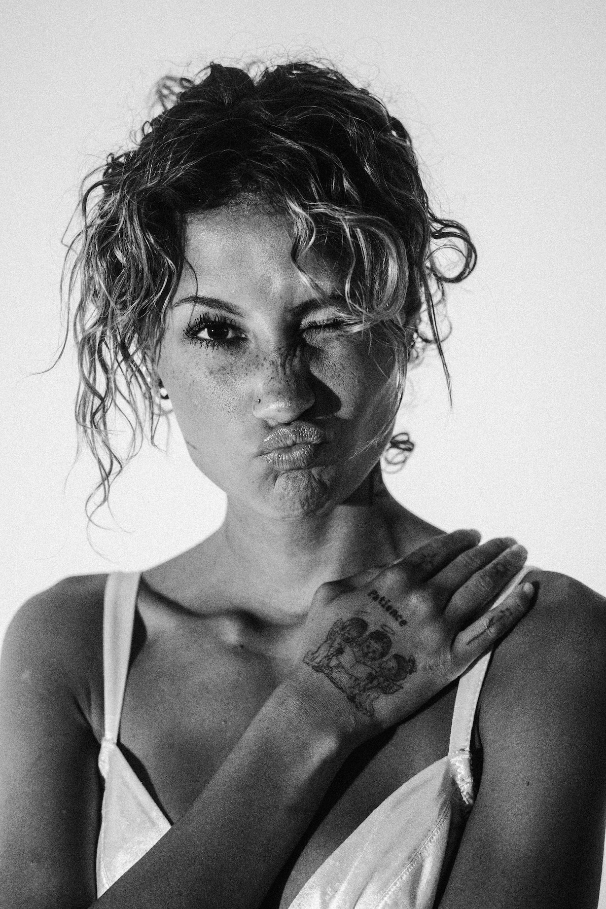 Close-up black and white portrait of a winking adult female with curly hair and a tattoo on her hand, against a plain white background.