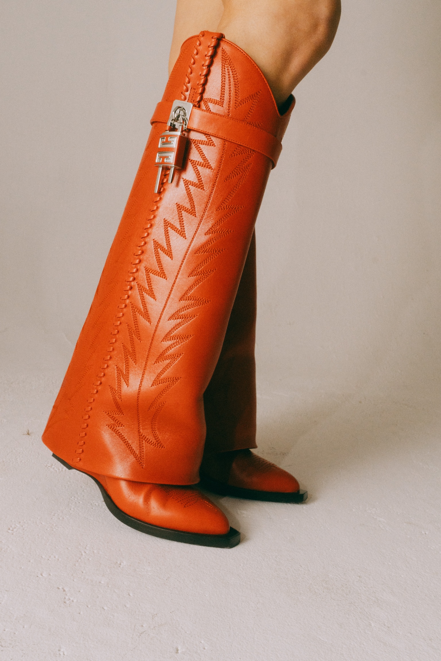 Fashionable orange high boots with decorative stitching, buckle, and silver hardware on light background in a studio setting.