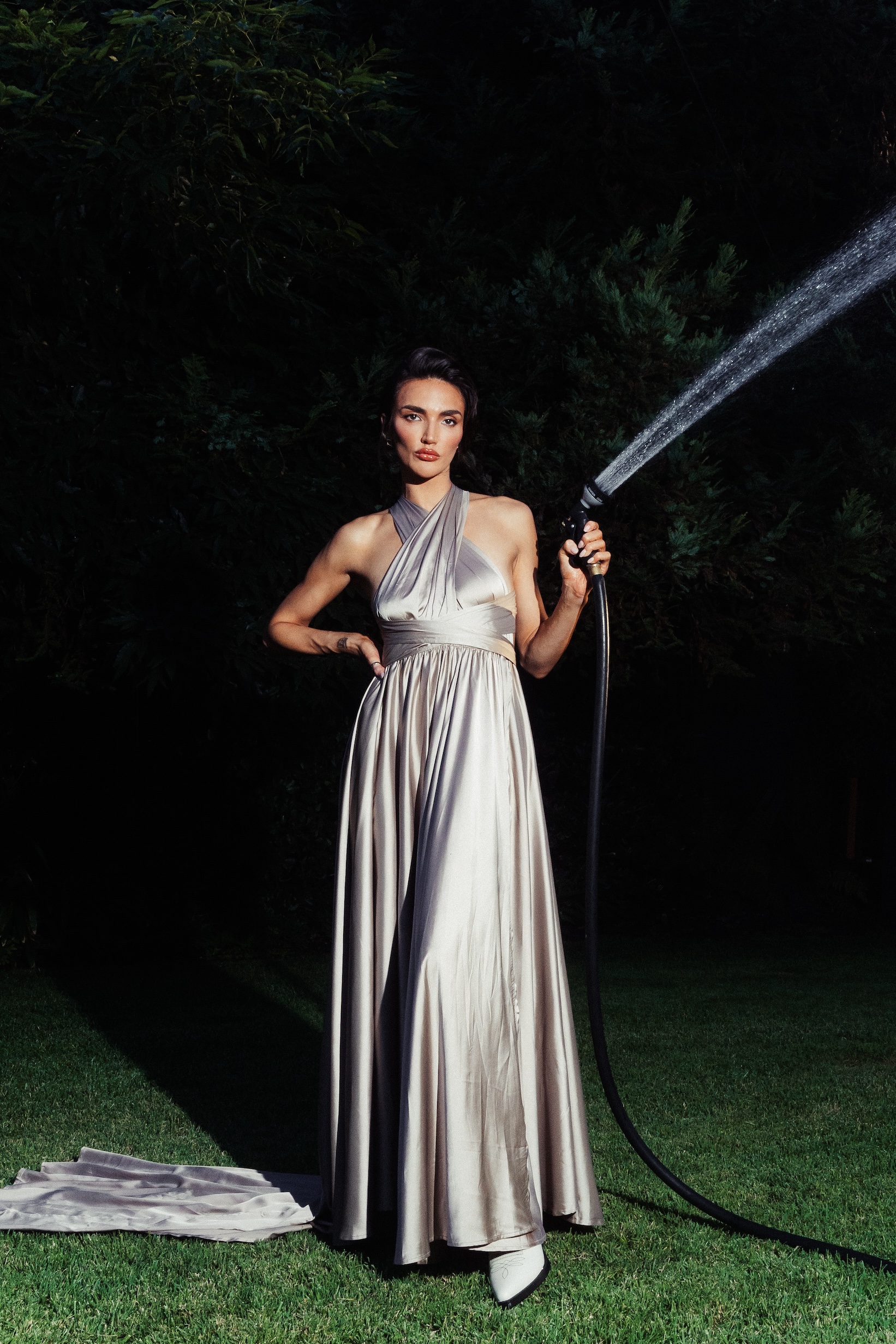 A woman in a long gown stands on a lawn holding a water hose, with water spraying against a backdrop of green foliage and a dark sky.