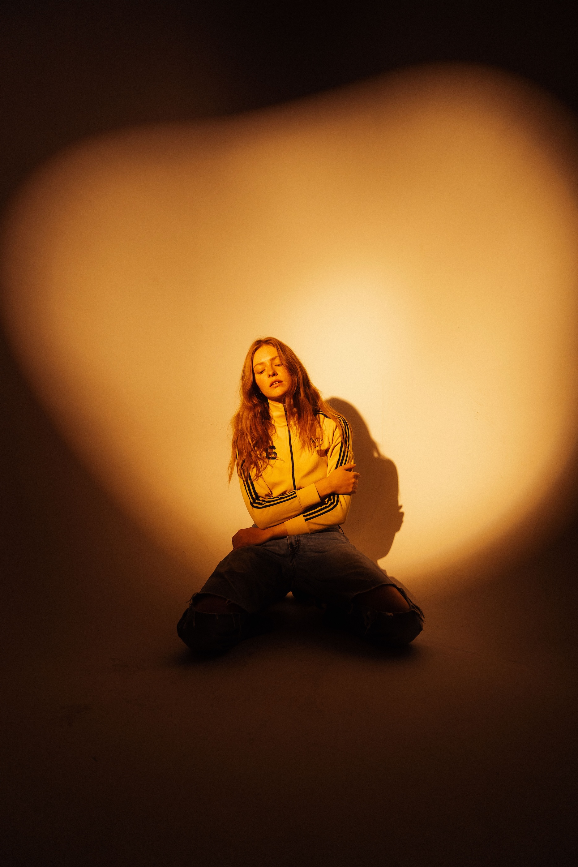 Young woman with long hair in a yellow Adidas track jacket and jeans posing indoors, with a dramatic spotlight and soft background lighting.