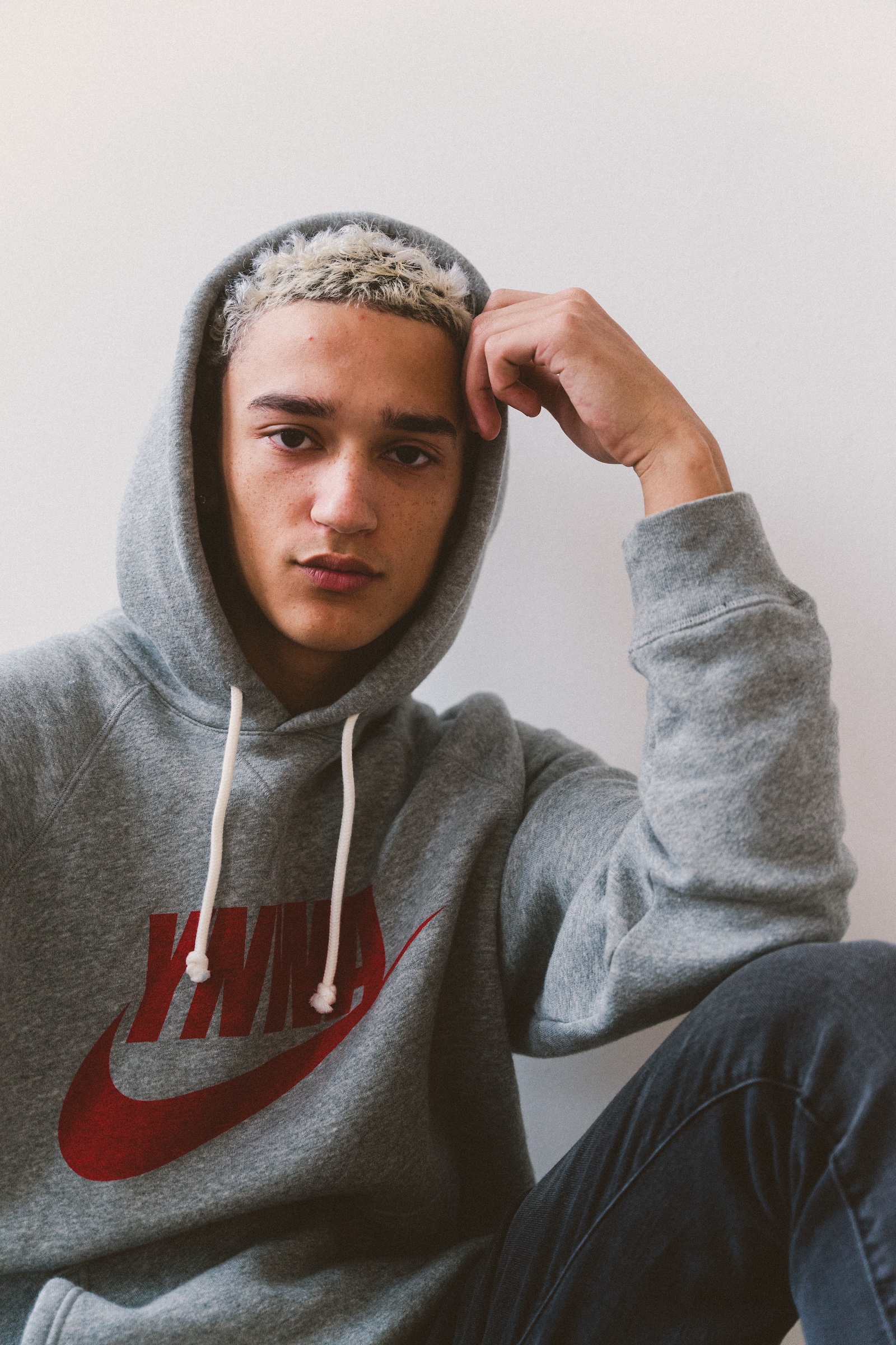 Portrait of a young adult with short blonde hair wearing a gray Nike hoodie and jeans sitting against a white wall indoors.