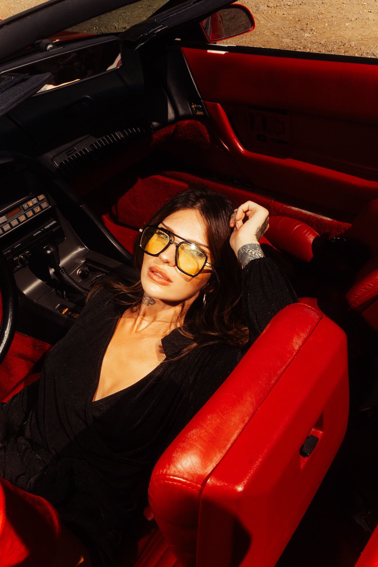 Brunette woman with yellow sunglasses reclines in red-leather interior of a classic black convertible car with tan desert backdrop.