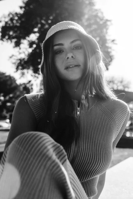 Black and white portrait of a woman wearing a bucket hat and a ribbed top, sitting outdoors with trees in the background.