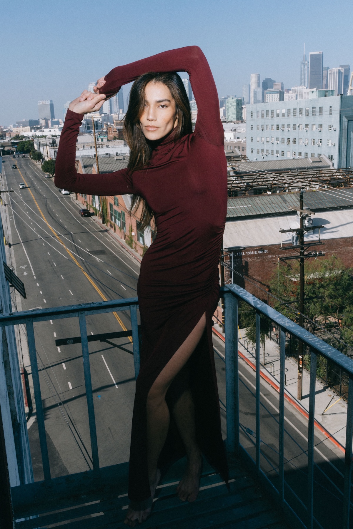 A brunette woman poses in a long maroon dress with a high slit against a cityscape backdrop, on a rooftop, under a clear blue sky.