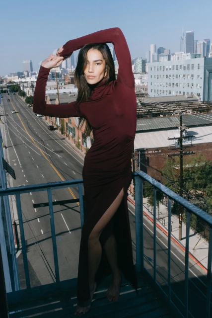 A brunette woman poses in a long maroon dress with a high slit against a cityscape backdrop, on a rooftop, under a clear blue sky.