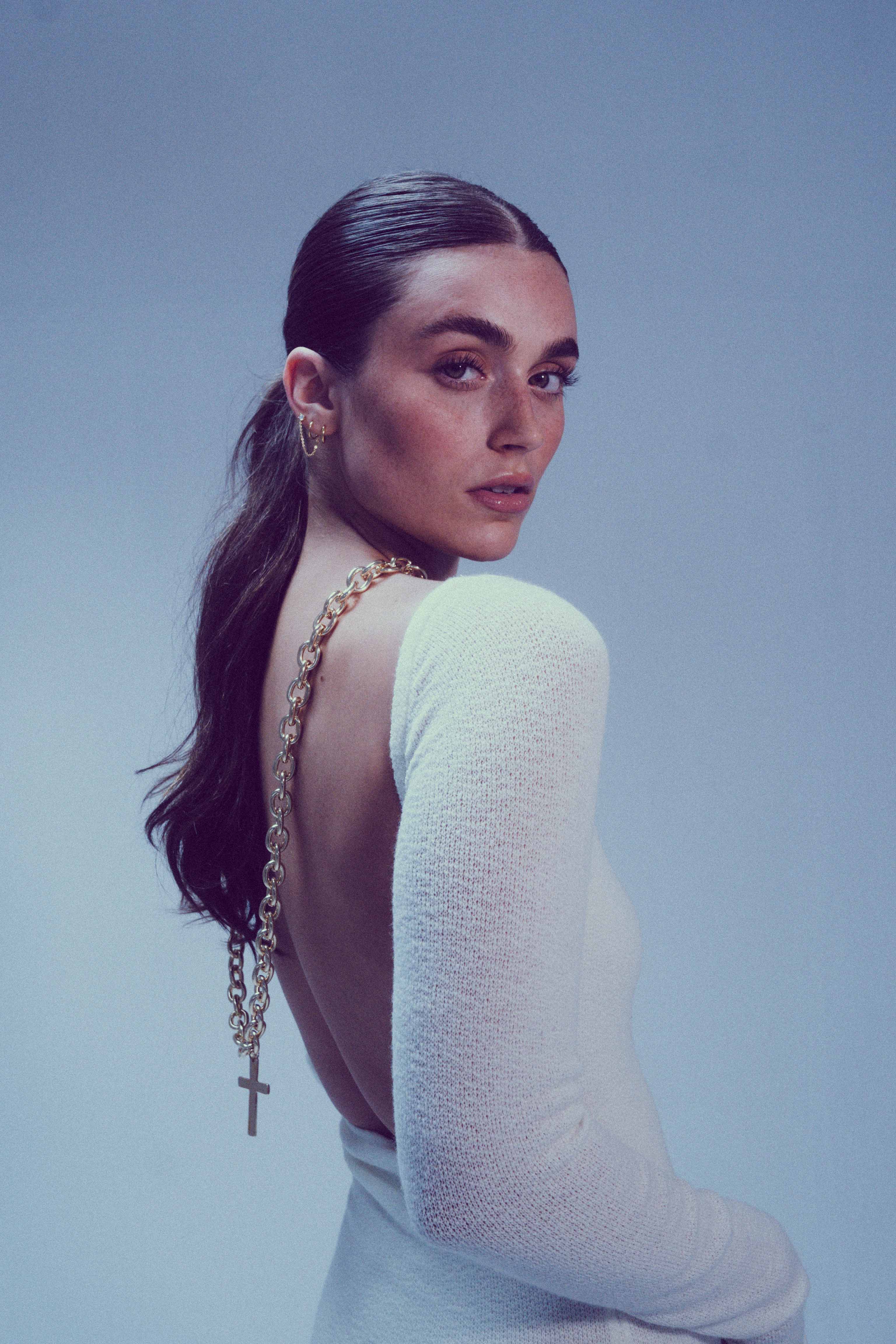 Portrait of a woman with dark hair pulled back in a ponytail, wearing a white dress and a long gold chain with a cross pendant.