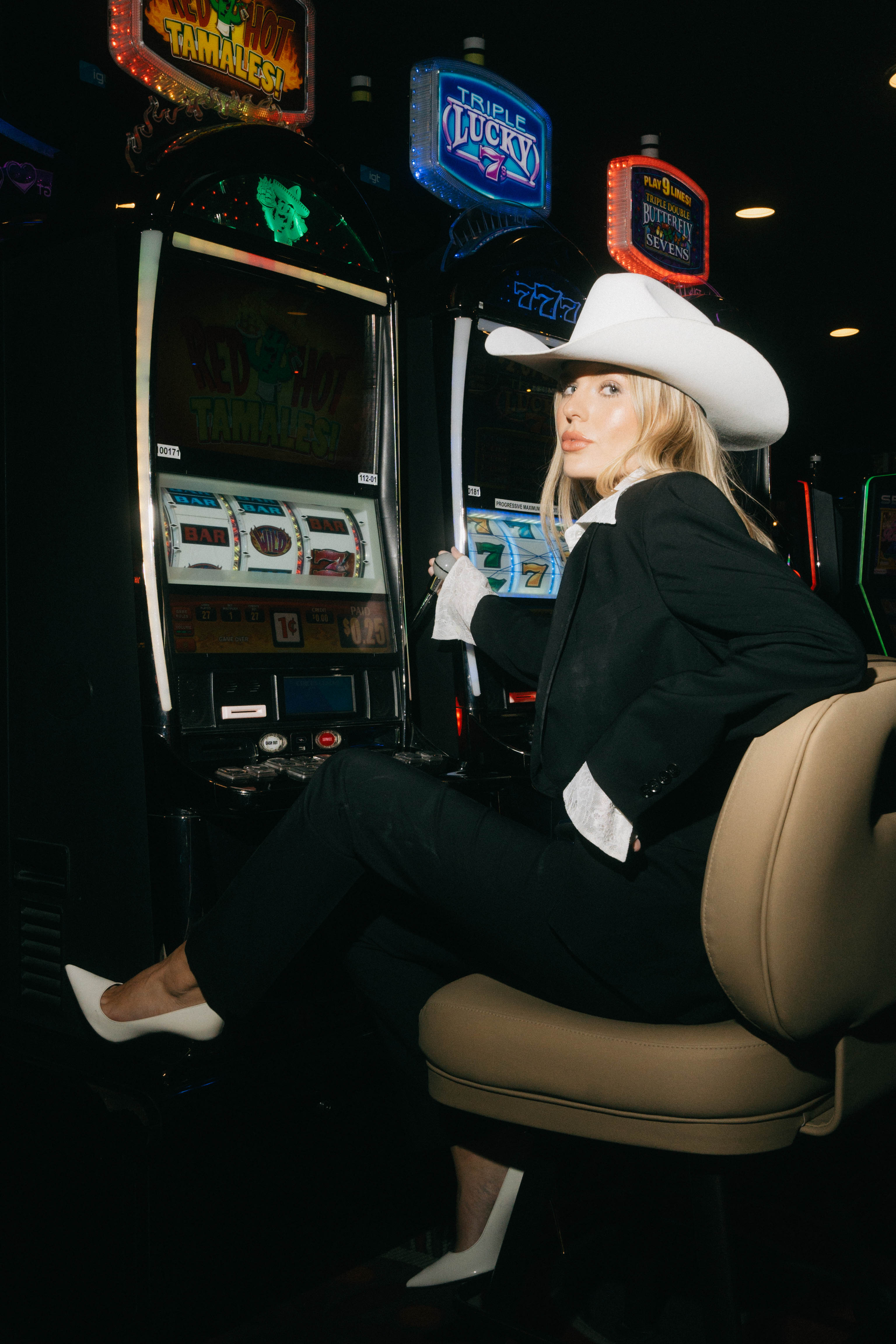 A woman in a black suit and white cowboy hat sits in a casino, posing in front of slot machines with bright, colorful lights.