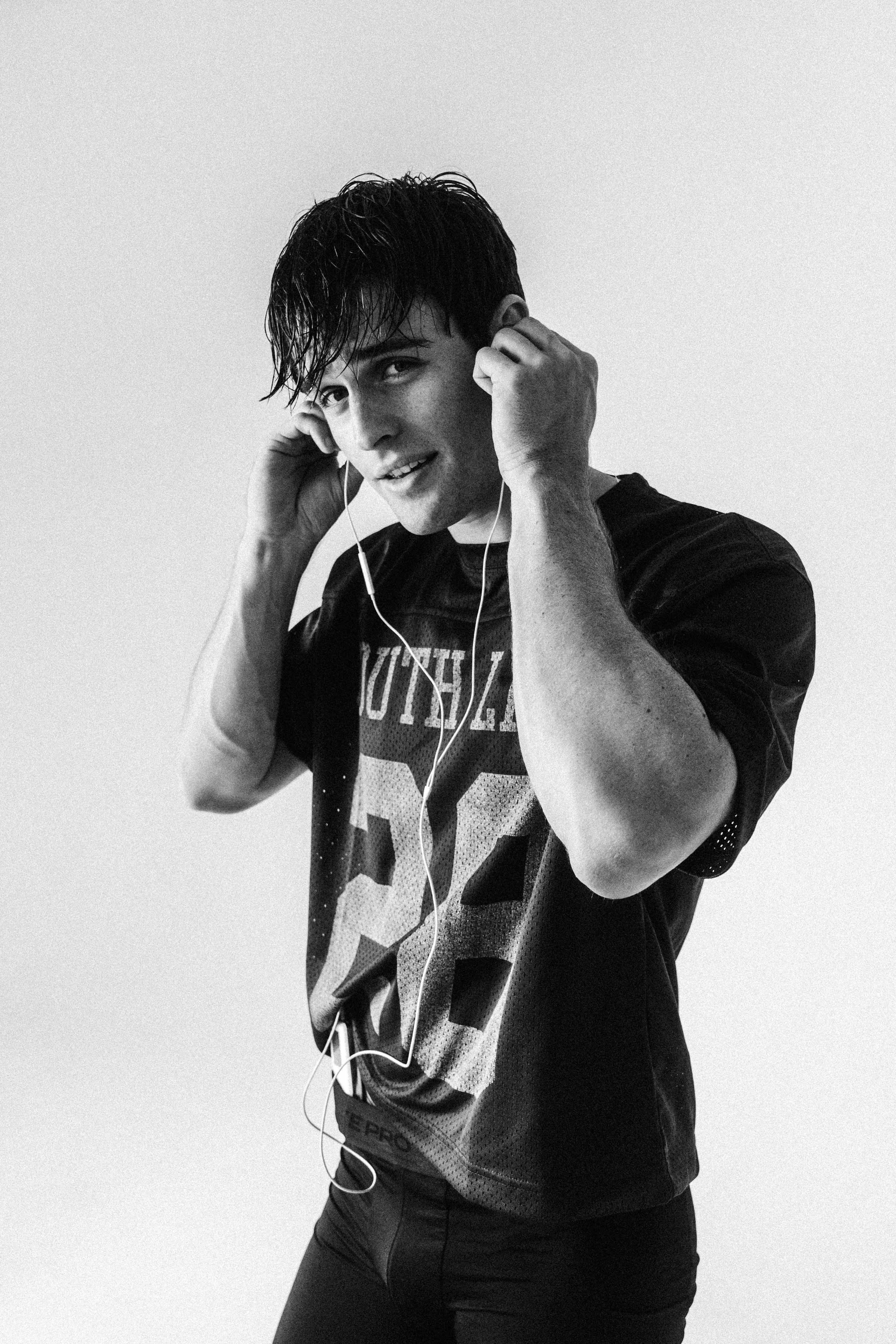 Black-and-white portrait of a young adult man wearing headphones, a football jersey, and leggings standing indoors with a neutral expression.