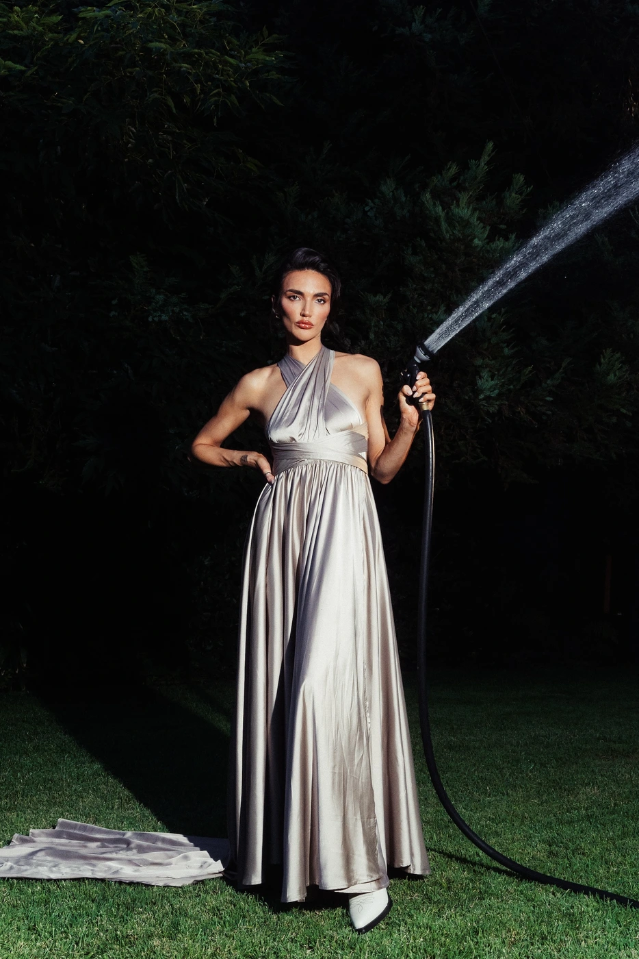 Woman in a silver satin halter gown standing on grass at night while spraying water from a garden hose.