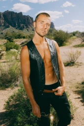 Man wearing a black leather vest and trousers in an arid desert landscape with mountains under a clear sky.