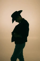 Silhouette of a man wearing a black cowboy hat and denim jacket looking down against a warm beige studio backdrop.