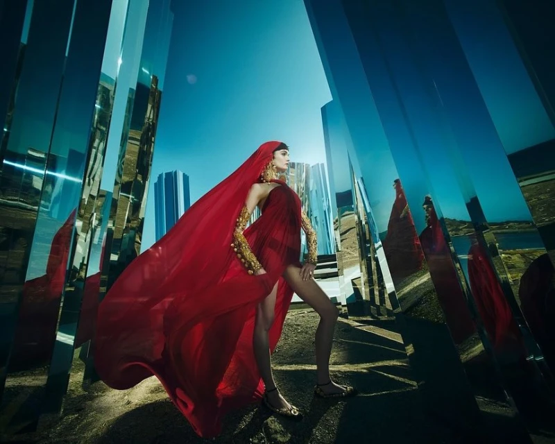 A woman poses in a flowing red dress in a stylized outdoor environment with mirrored pillars and a clear blue sky.