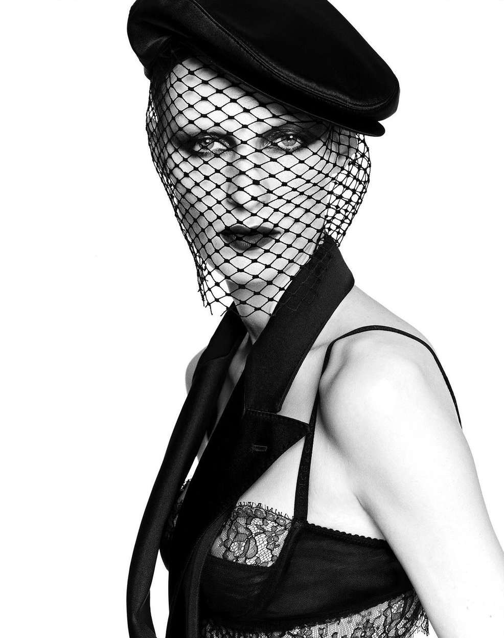 Black-and-white portrait of a woman wearing a cap and veil, gazing intensely at the camera in a photo studio setting with a minimalist background.