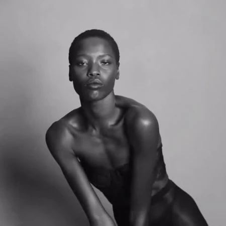 Portrait of a black woman with short hair in dark clothing, against a light grey background, demonstrating a sensual, confident pose.