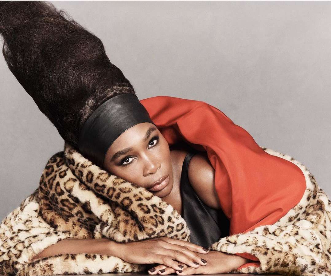 Portrait of a Black woman wearing a tall hairstyle and a leopard print coat with a red lining against a solid grey background.
