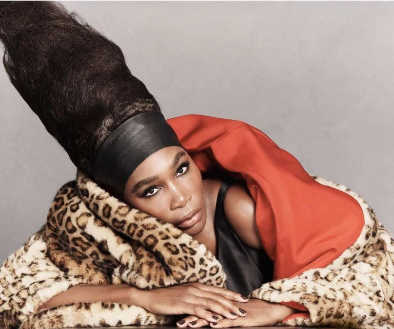 Portrait of a Black woman wearing a tall hairstyle and a leopard print coat with a red lining against a solid grey background.
