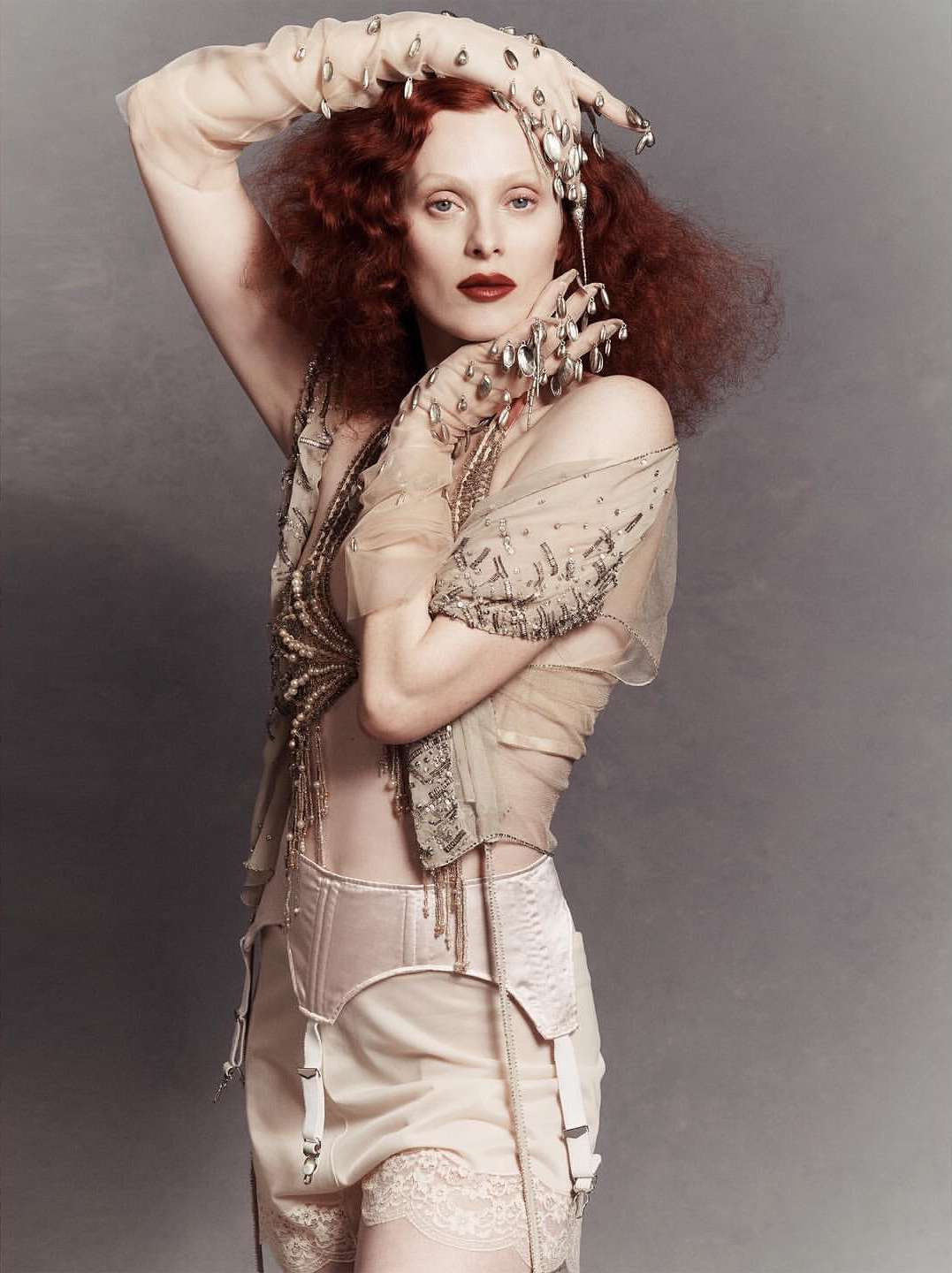 A pale-skinned woman with curly red hair poses in lingerie and sheer gloves, creating a vintage fashion editorial look against a gray backdrop.
