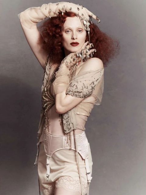 A pale-skinned woman with curly red hair poses in lingerie and sheer gloves, creating a vintage fashion editorial look against a gray backdrop.