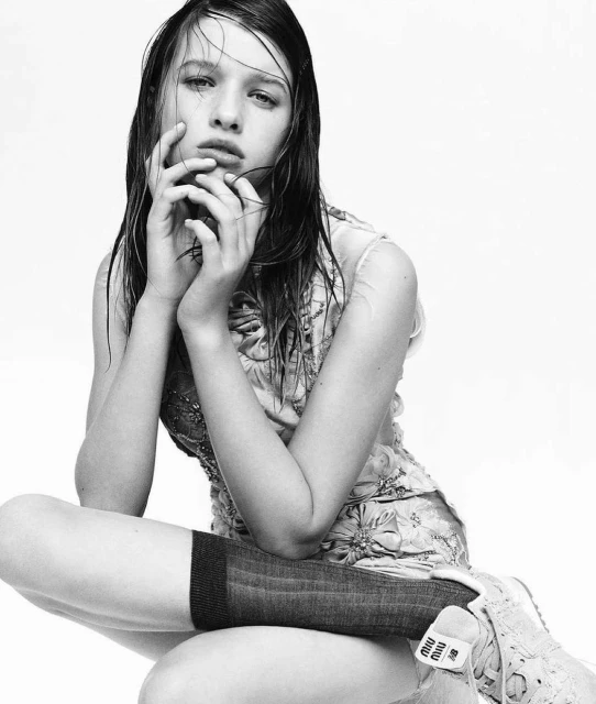 Black and white portrait of a young woman with wet dark hair and a floral top, sitting with hands up to her face in a studio setting.