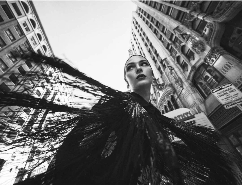 Monochrome fashion shot of a woman in an ornate shawl, posing in an urban environment with tall buildings under a bright sky.