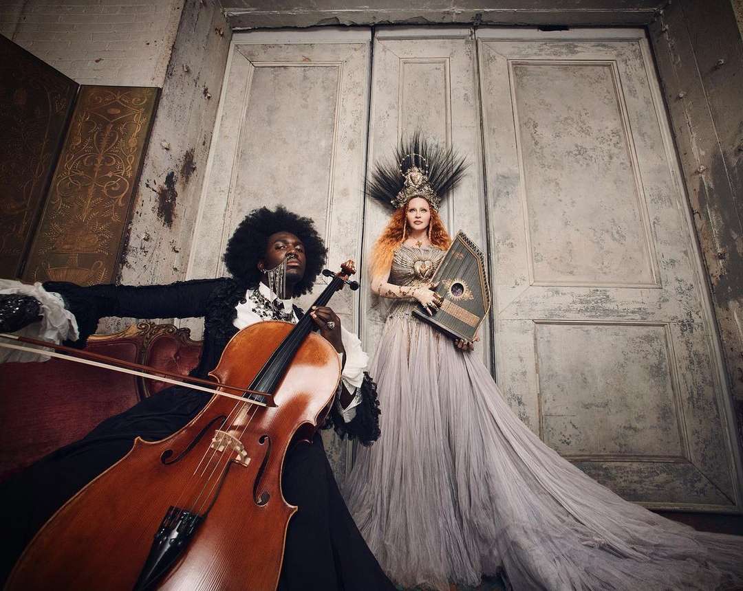 Musical performers pose indoors with cello and harp in ornate gowns against weathered walls and doors in dramatic, soft lighting.