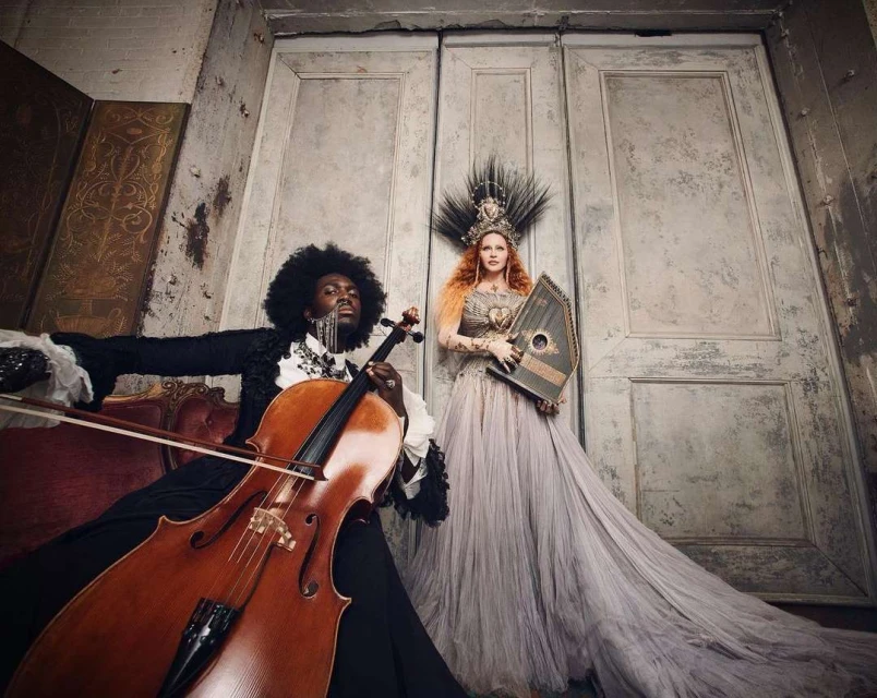 Musical performers pose indoors with cello and harp in ornate gowns against weathered walls and doors in dramatic, soft lighting.