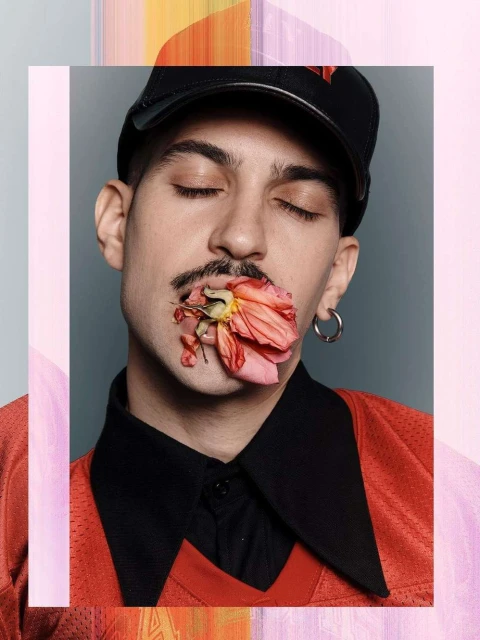 Portrait of a young adult with a mustache, wearing a cap and a red jersey, with a pink flower in their mouth, set against a grey background.