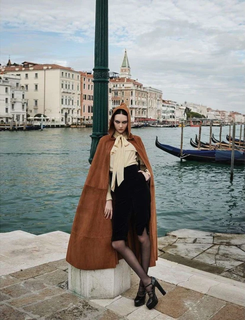 Fashion shot of an adult female model wearing a cape and heels, posing on a stone walkway with canals and buildings in the background.