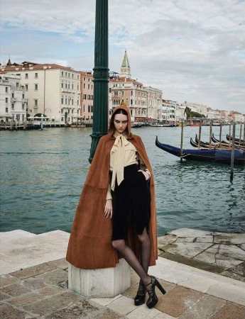 Fashion shot of an adult female model wearing a cape and heels, posing on a stone walkway with canals and buildings in the background.