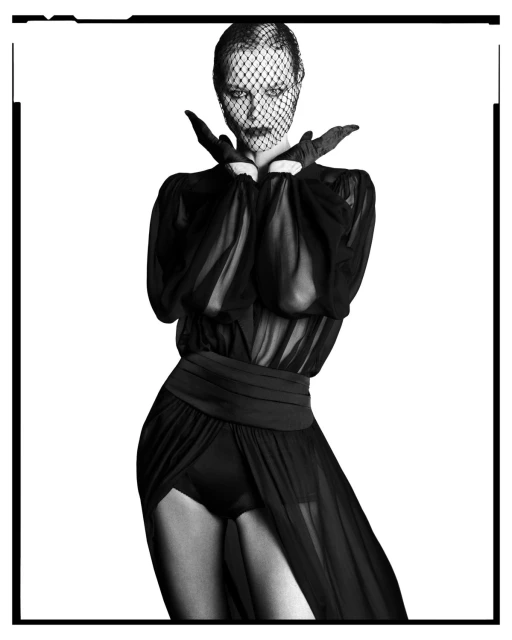 Monochrome fashion portrait of an adult female wearing a black veil, gloves, and a sheer dress against a stark white backdrop.