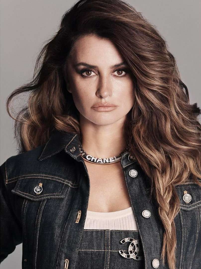 Close-up portrait of adult woman with long brown hair, wearing a dark denim jacket and Chanel necklace in a photo studio setting.