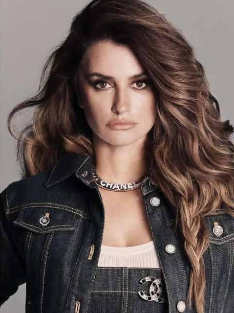 Close-up portrait of adult woman with long brown hair, wearing a dark denim jacket and Chanel necklace in a photo studio setting.