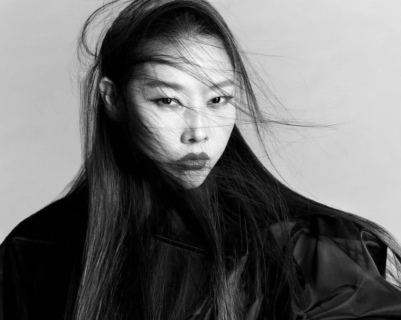 Monochrome portrait of an East Asian woman with long dark hair blowing across her face and wearing a dark coat against a plain background.