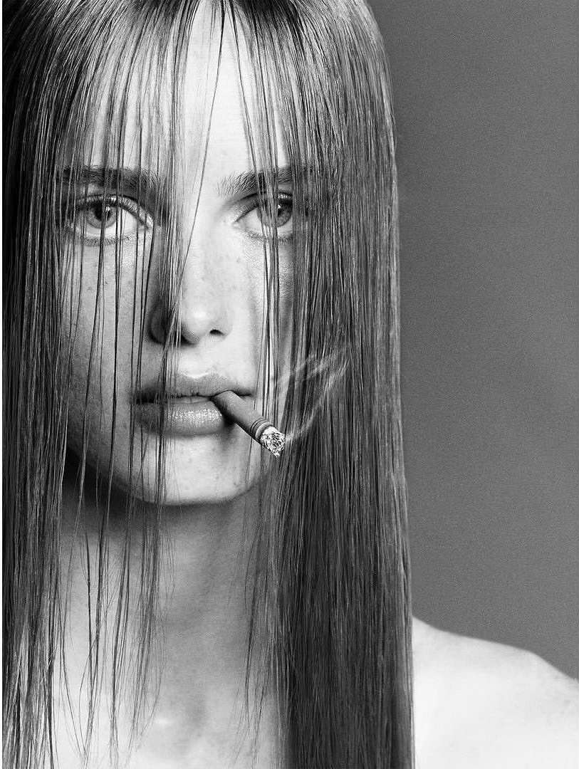 Monochrome portrait of a young adult woman with long hair, cigarette in mouth, and smoke around face against gray background.