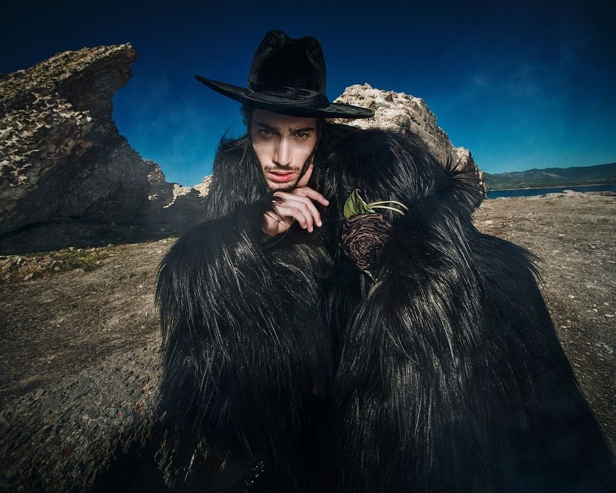 A male model in a black hat and furry coat poses outdoors against a rocky landscape with a distant body of water under a blue sky.