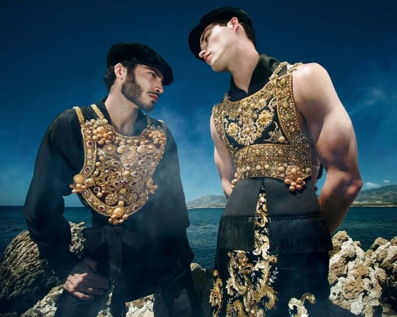 Two men wearing black outfits with ornate gold embroidery and caps posing against a coastal rocky landscape under a blue sky.