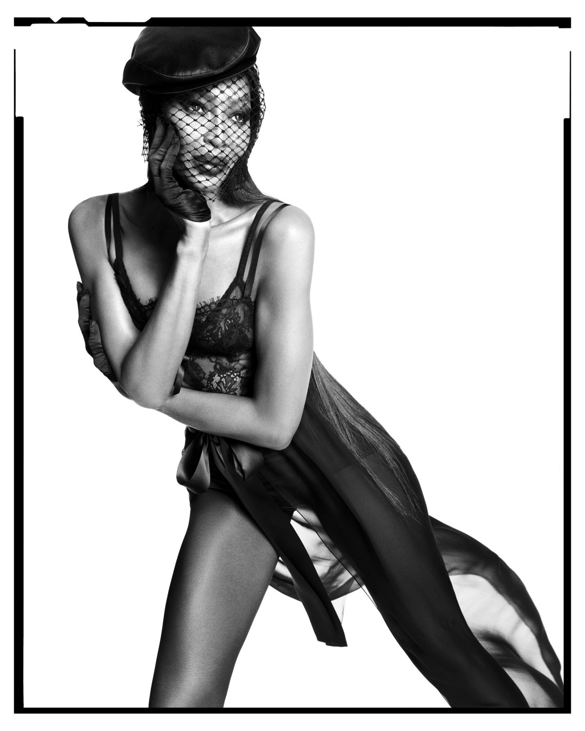 Black and white portrait of a woman wearing a hat and veil, striking a pose in lingerie and a sheer robe, against a white backdrop.