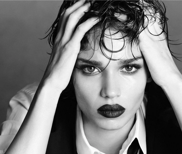 Close-up black and white portrait of a woman with wet hair, dark lipstick, wearing a shirt and vest, against a neutral background.